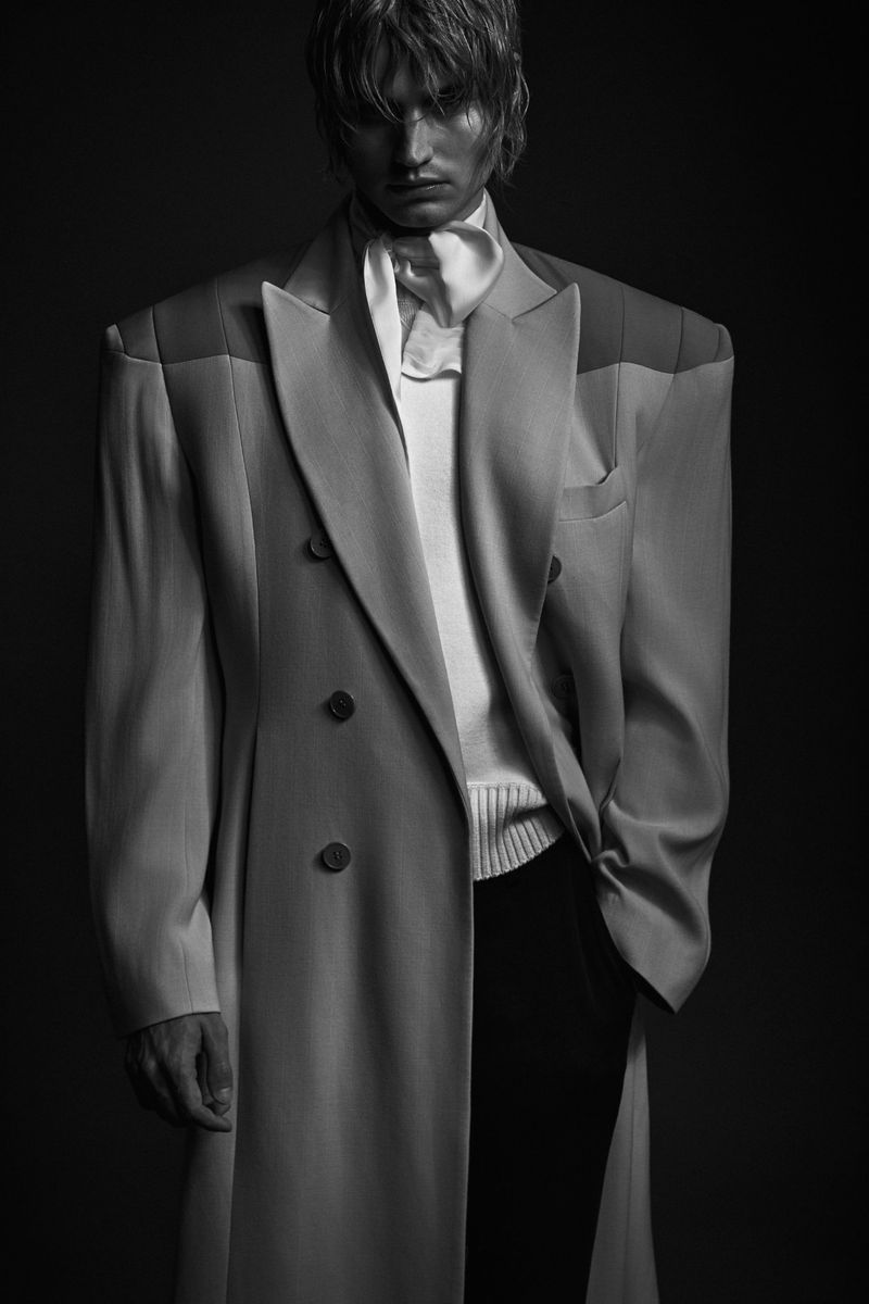 Monochrome fashion shot of a blonde adult male wearing a long gray coat, white top, and dark trousers, in a dark photo studio.
