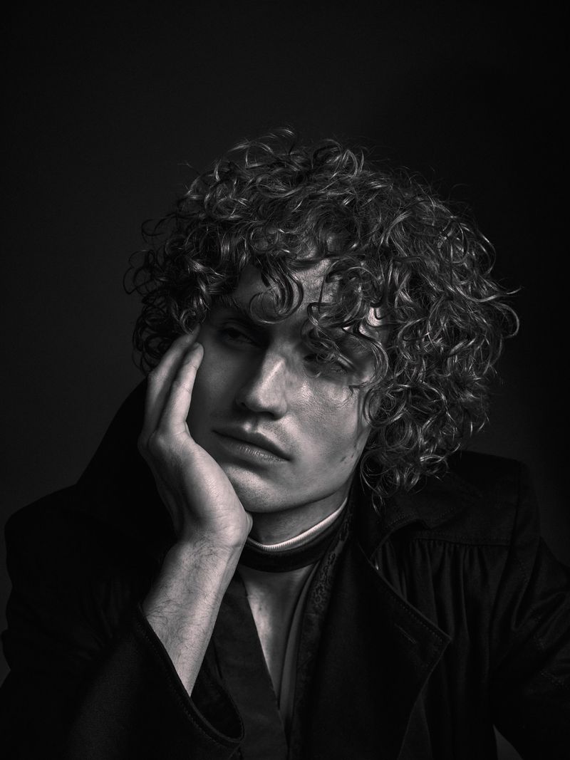 Black and white portrait of a curly-haired man with his hand to his face, wearing a dark jacket, against a dark background.