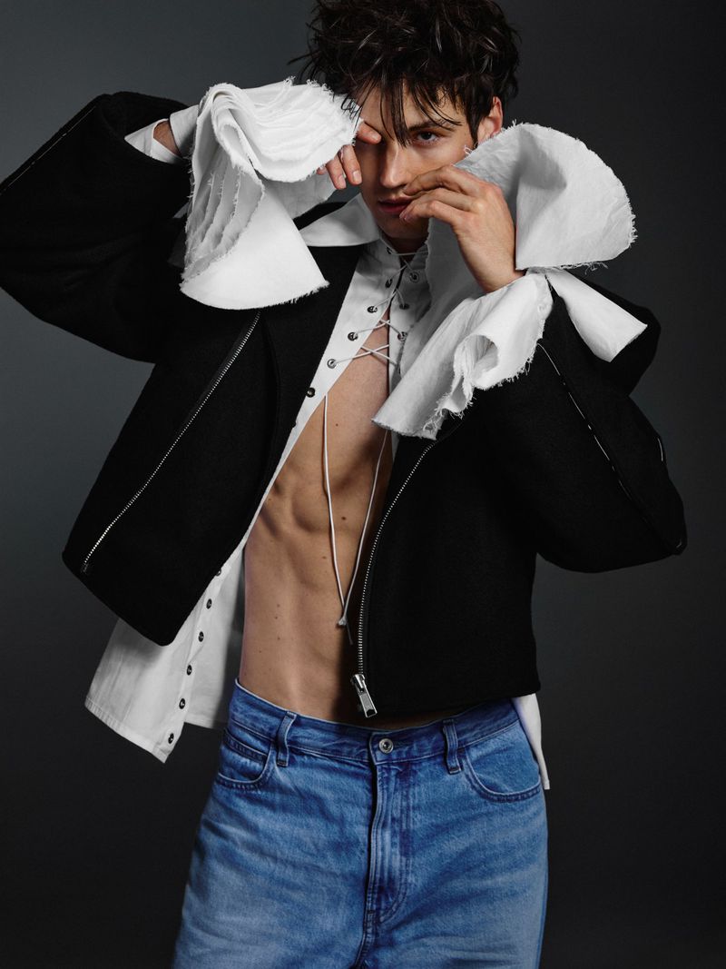 Male model wearing a black jacket, laced white shirt and blue denim jeans against a dark grey background.