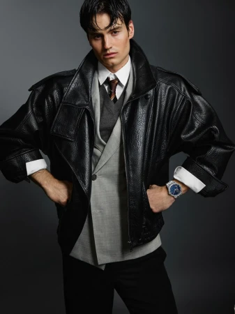 Handsome male model poses in a gray suit, black leather jacket, tie, and black pants against a solid gray backdrop in a fashion editorial.