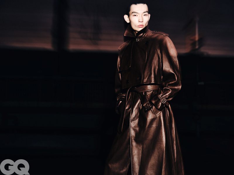 Male model wearing a long, brown leather trench coat in a studio environment with a dark background and the GQ logo visible.