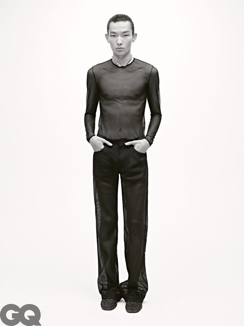 Full-length portrait of an Asian male fashion model in black see-through long-sleeved shirt and semi-sheer pants on a white background.