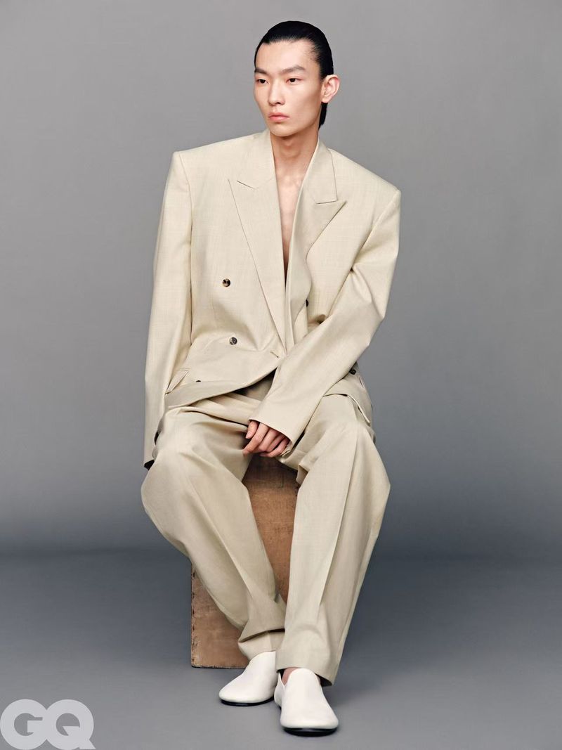 Adult male posing in an oversized beige suit and white slip-on shoes, seated on a wooden cube against a grey studio backdrop.