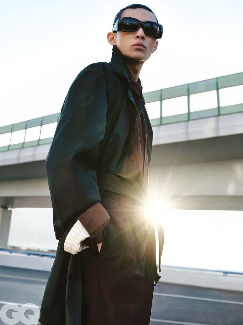 An Asian male model stands on an overpass wearing a black coat and dark sunglasses against a bright sky with sun glare.