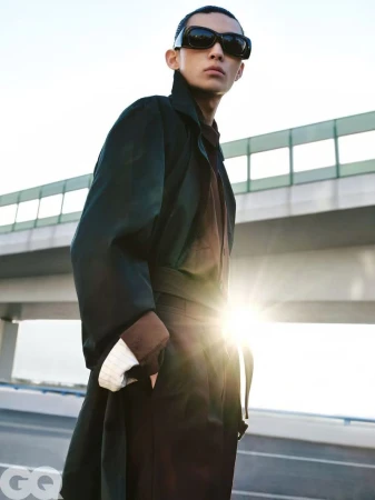 An Asian male model stands on an overpass wearing a black coat and dark sunglasses against a bright sky with sun glare.