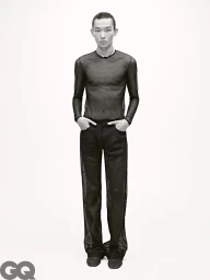 Model wearing a sheer black long-sleeve top and matching trousers with hands in pockets against a plain white background.