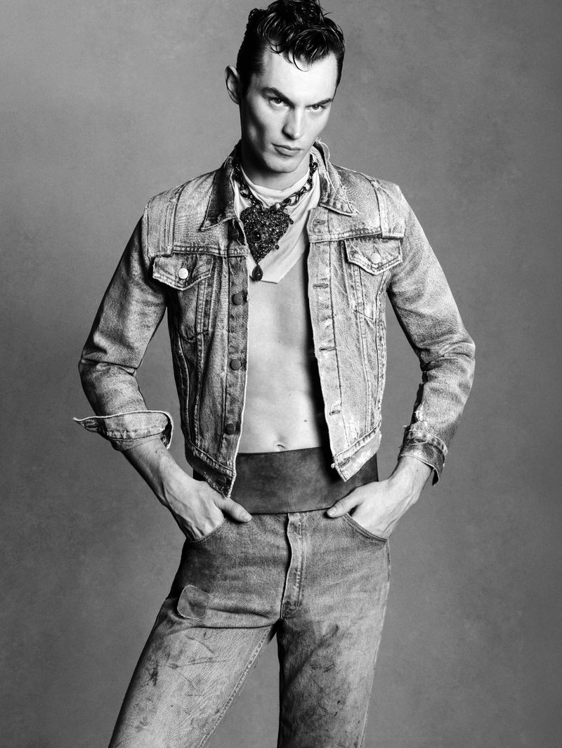 Black and white portrait of a male fashion model with a denim jacket, jewelry, and jeans in a photo studio with a grey background.