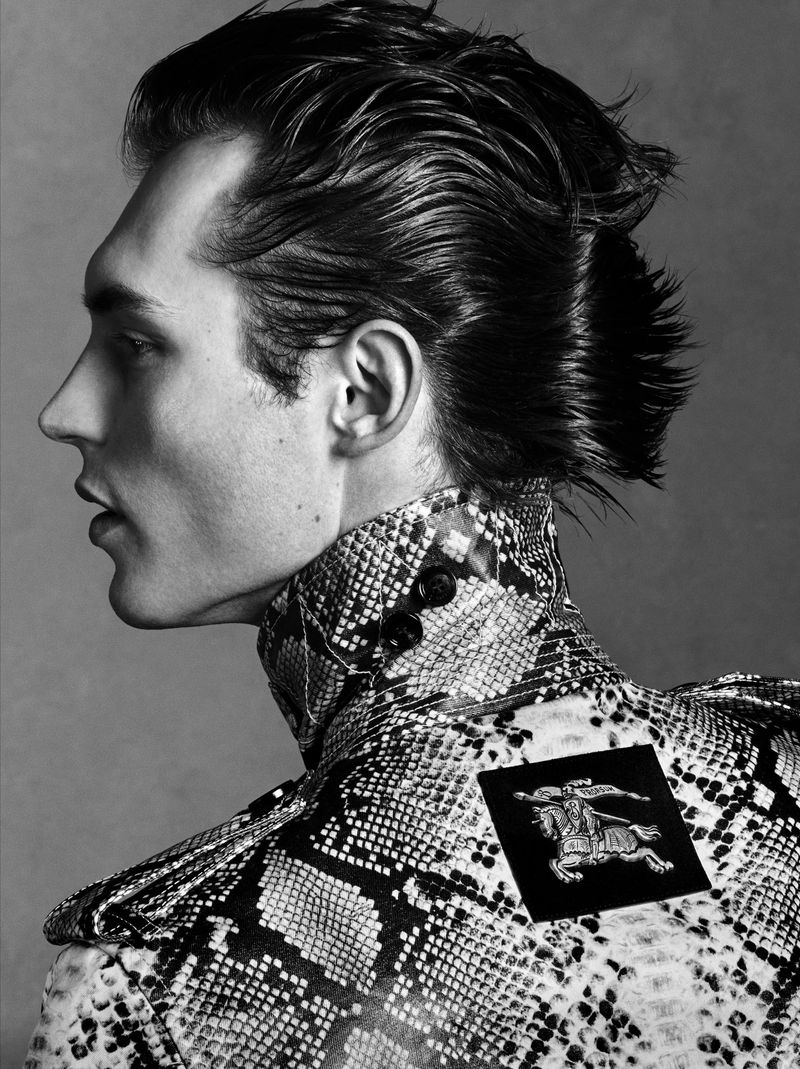 Black and white portrait of a male fashion model with dark hair wearing a snakeskin jacket with a high collar, against a grey background.