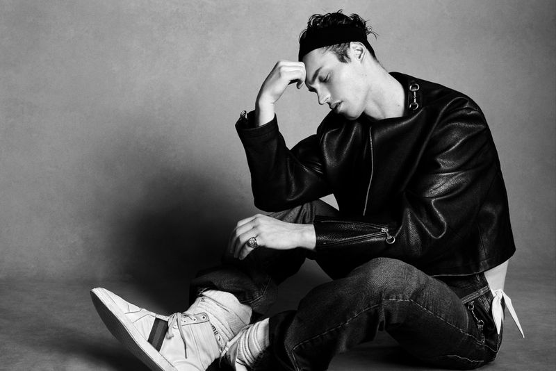 Kit Butler sitting on the floor in a black leather jacket, dark jeans, and white sneakers against a neutral grey studio backdrop.