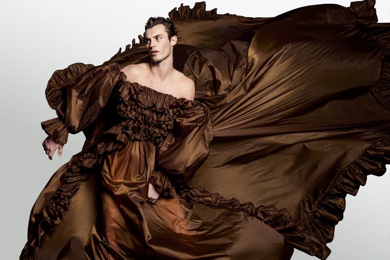 A male fashion model poses in a billowing, off-the-shoulder brown gown with ruffled trim, against a neutral background, studio lighting.