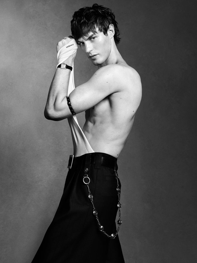 Black and white fashion shot of a muscular adult male wearing black pants with chains, posing shirtless with a white fabric wrap.