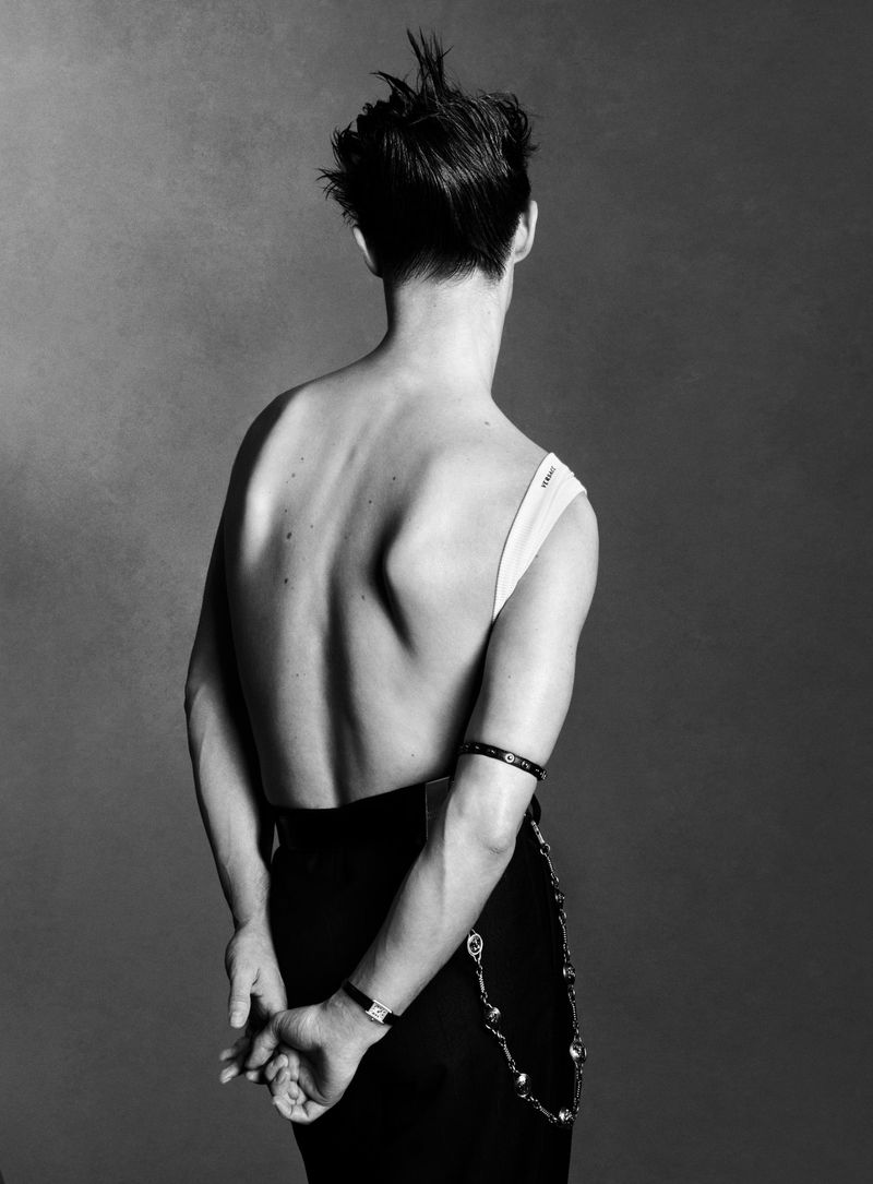 Back view of a shirtless adult man wearing a skirt and jewelry posing in a studio with a gray background in a black and white photography.