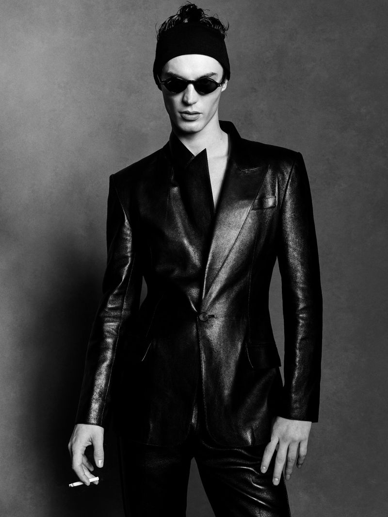 Monochrome studio portrait of a male fashion model in a leather suit, beanie, and sunglasses, holding a cigarette, against a grey backdrop.