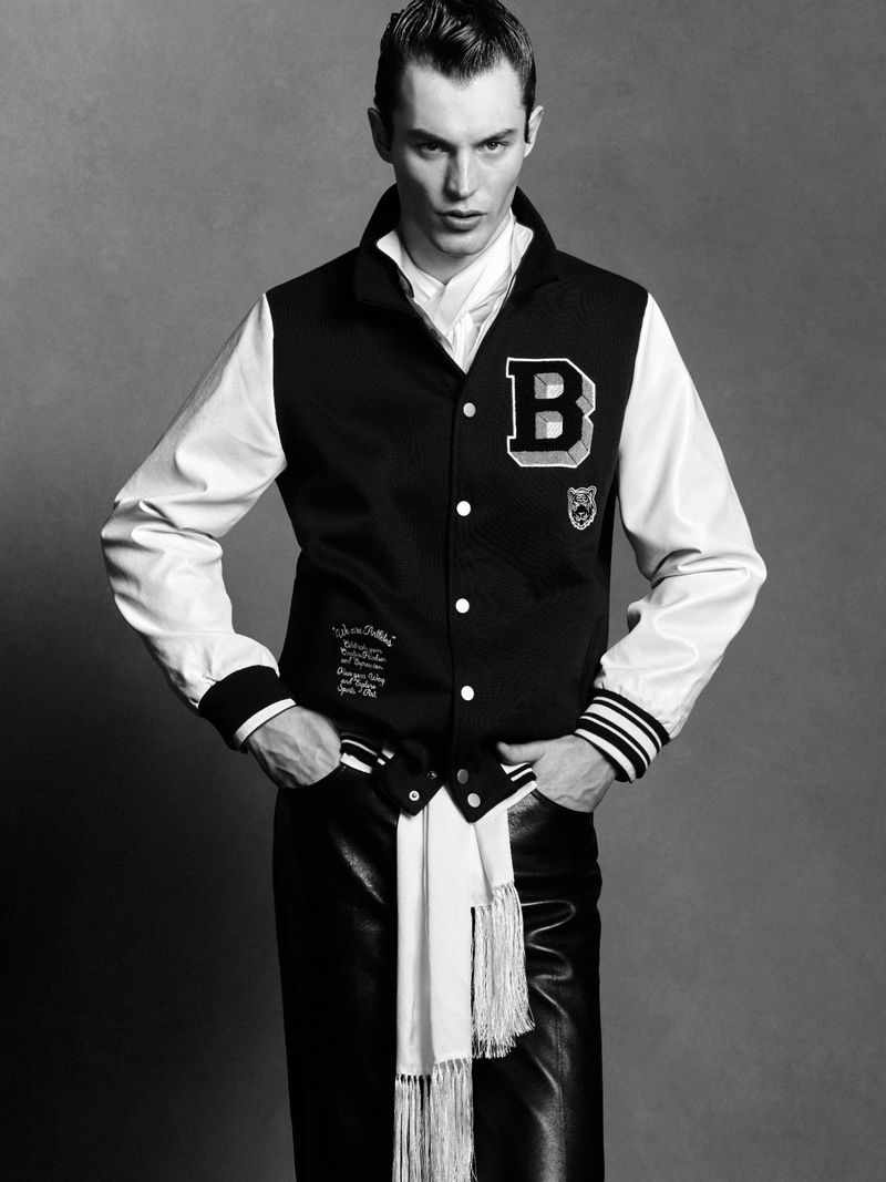 Black and white portrait of a male fashion model wearing a letterman jacket and leather pants against a grey backdrop in a studio.