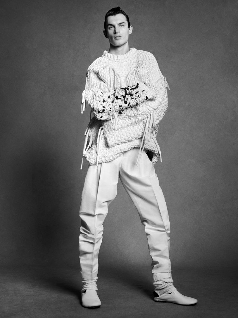 Black and white studio portrait of a male fashion model wearing a sweater and trousers, posing against a grey backdrop.