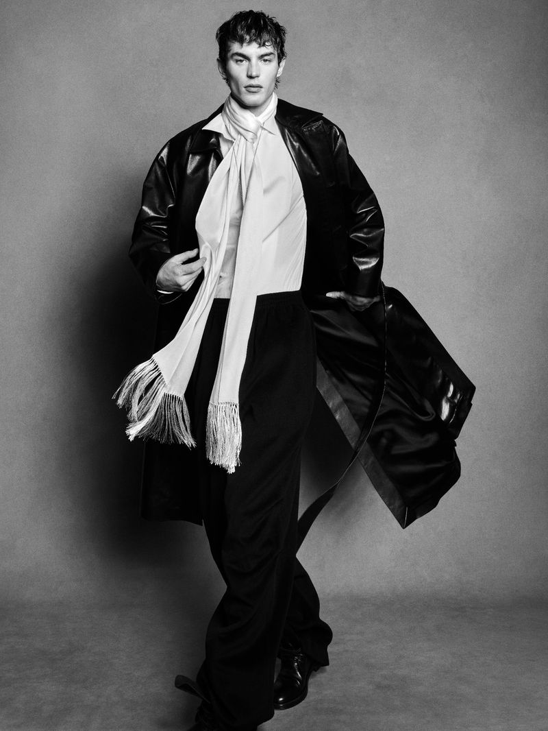 Black and white portrait of a young adult male fashion model in a black overcoat, wide-leg trousers, and a white scarf, posing in a studio.