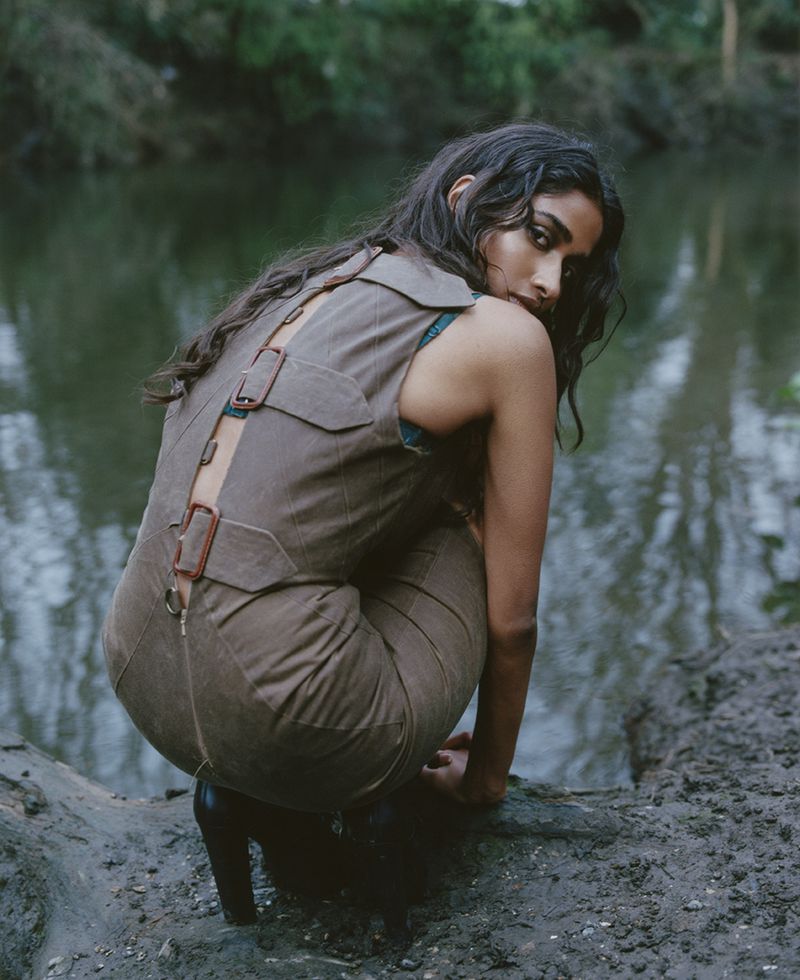 Woman with dark hair poses in a brown jumpsuit by a murky river. Fashion shot with lush forest background and soft, natural lighting.