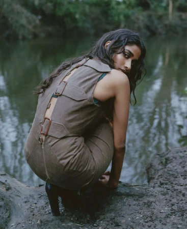 Woman with dark hair poses in a brown jumpsuit by a murky river. Fashion shot with lush forest background and soft, natural lighting.
