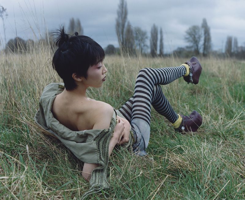 Asian woman lies in tall grass wearing striped leggings, yellow socks and dark shoes, along with a green jacket and hair in a top knot.
