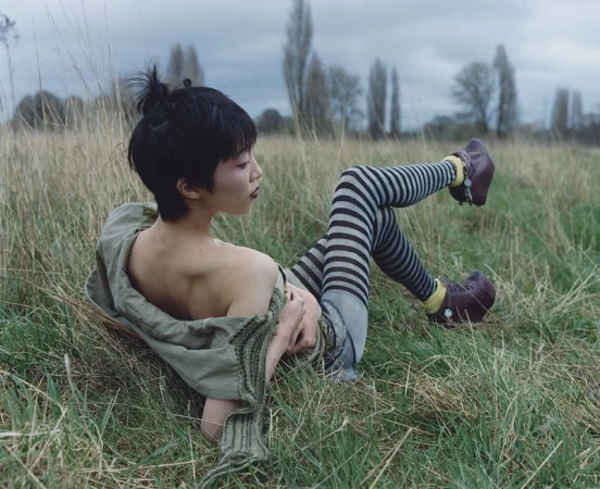 Asian woman lies in tall grass wearing striped leggings, yellow socks and dark shoes, along with a green jacket and hair in a top knot.