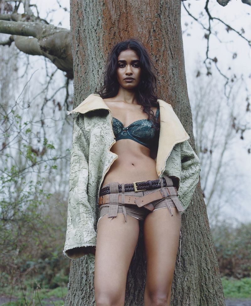 Adult woman posing outdoors in underwear and jacket leaning against a tree in a wooded area during daytime with natural lighting.