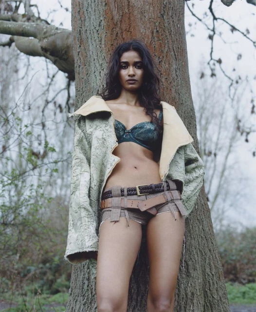 Adult woman posing outdoors in underwear and jacket leaning against a tree in a wooded area during daytime with natural lighting.