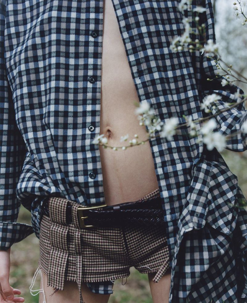 Unbuttoned plaid shirt, brown plaid shorts, black braided belt, and white flowers frame a woman outdoors in natural light.