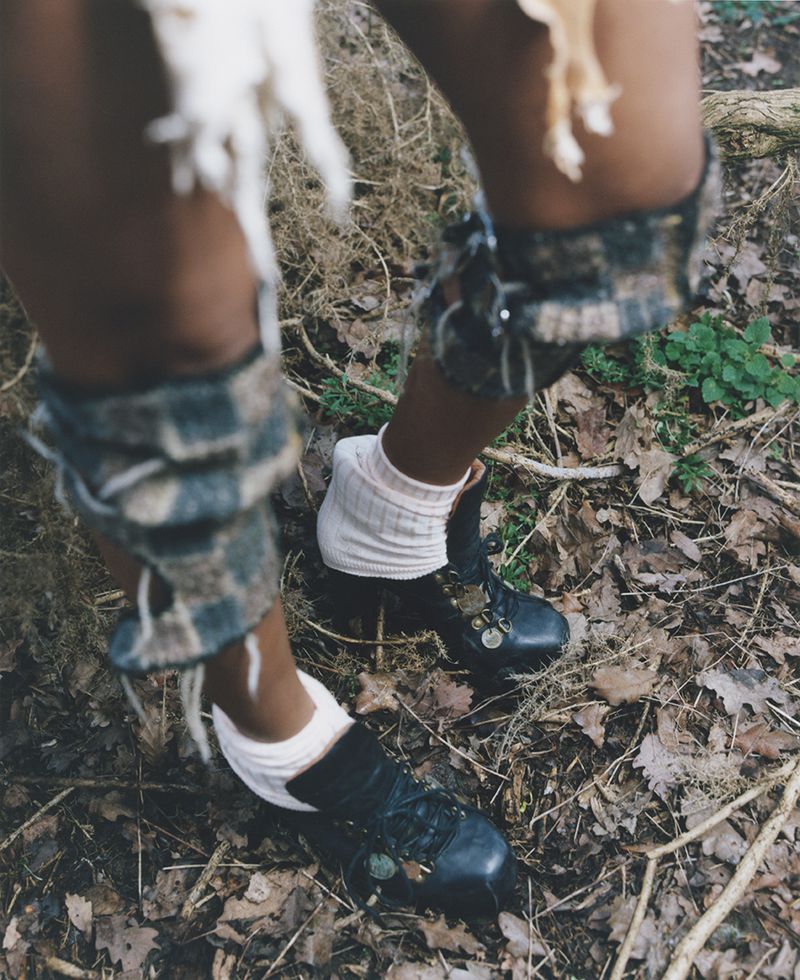 Close-up ground-level shot of a person's lower legs wearing white socks, black shoes, and plaid leg coverings, standing on fallen leaves.