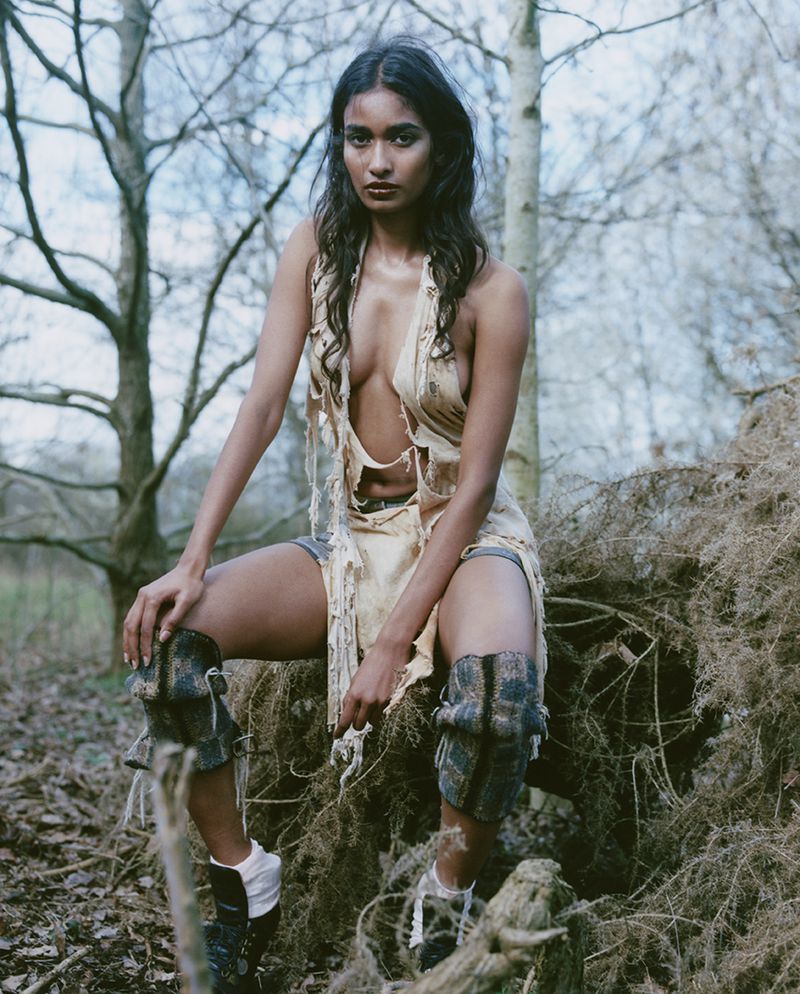 Woman in tattered dress and knee pads sitting on branches in a forest. Fashion editorial portrait featuring a female model. Outdoor setting.