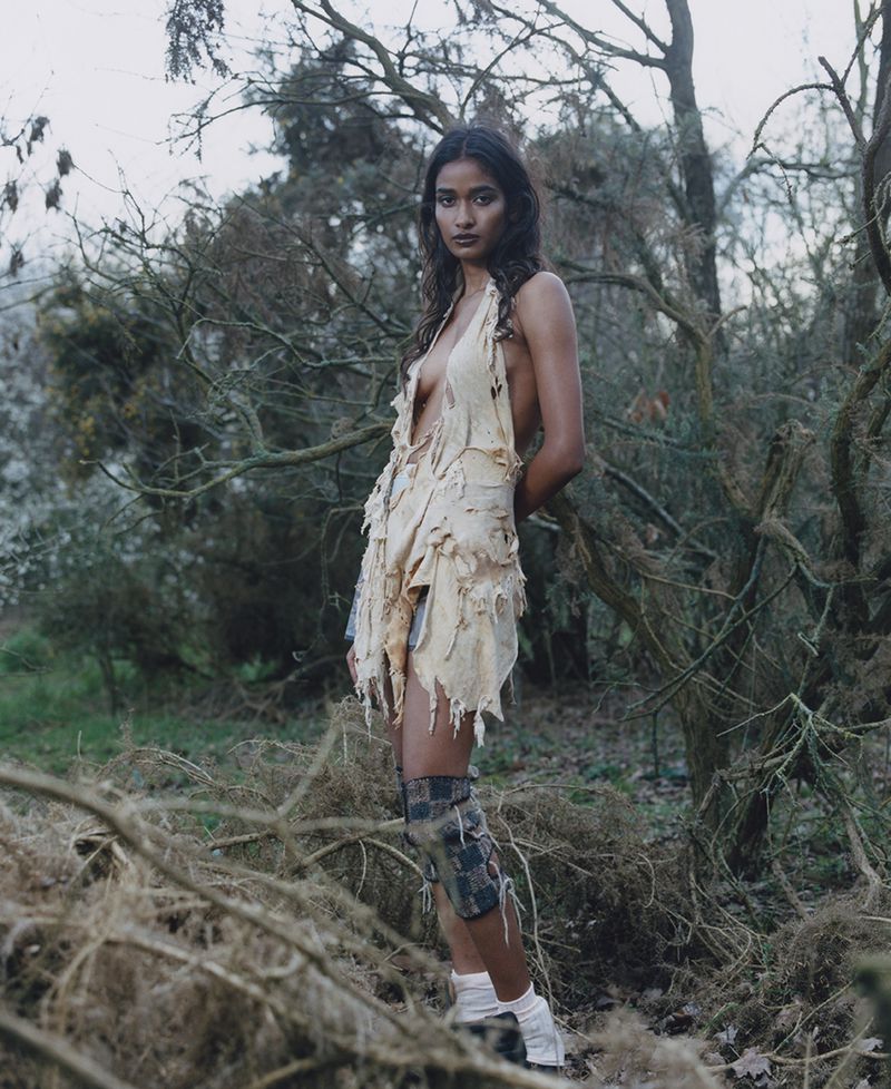 Portrait of a slender young woman with long dark hair in a tattered light yellow dress, knee pad, and white shoes, standing in a forest.