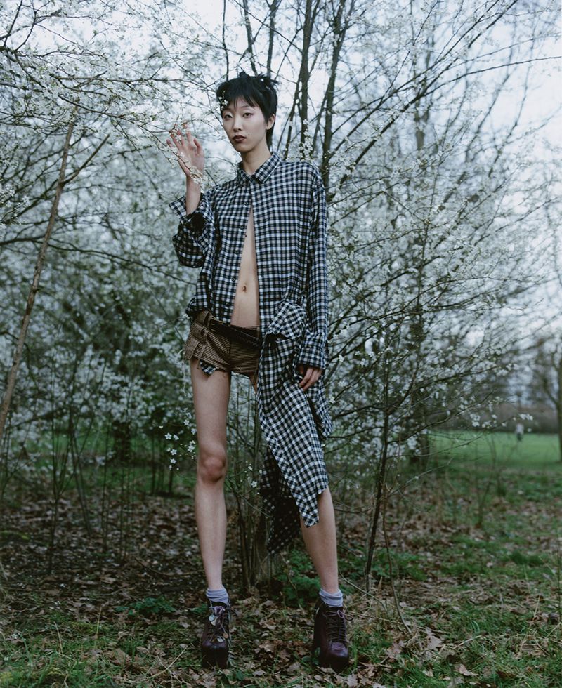 Asian woman in a plaid shirt and brown shorts stands among flowering trees in a grassy park. The scene has a natural, editorial feel.
