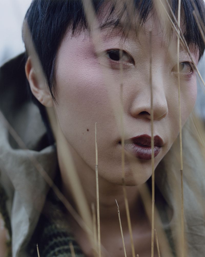 Close-up portrait of an Asian female with dark hair, pink blush, and dark lipstick, partially obscured by dried grass in a natural outdoor setting.