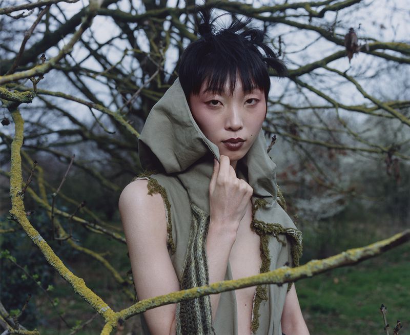 Asian woman with dark hair poses in an olive-green, hooded vest in front of bare branches for a fashion or beauty editorial.