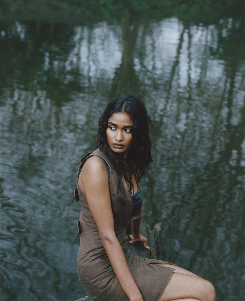 A woman with long dark hair wearing a brown dress sits on a log in front of water with trees reflected in it, captured in a medium shot.