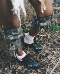 Close-up shot of model legs wearing knee wraps, white socks, and dark boots standing on a forest floor covered in dry leaves.