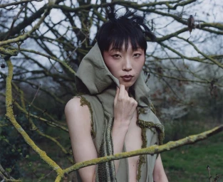 Model in a hooded green garment posing among bare branches in an outdoor setting