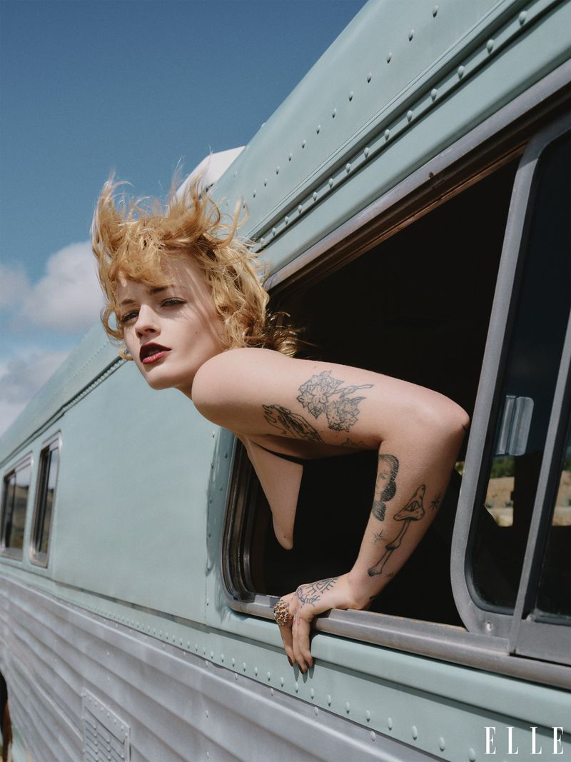 Adult female with arm tattoos leans out the window of a turquoise bus under a blue sky, magazine cover, medium shot, outdoor.