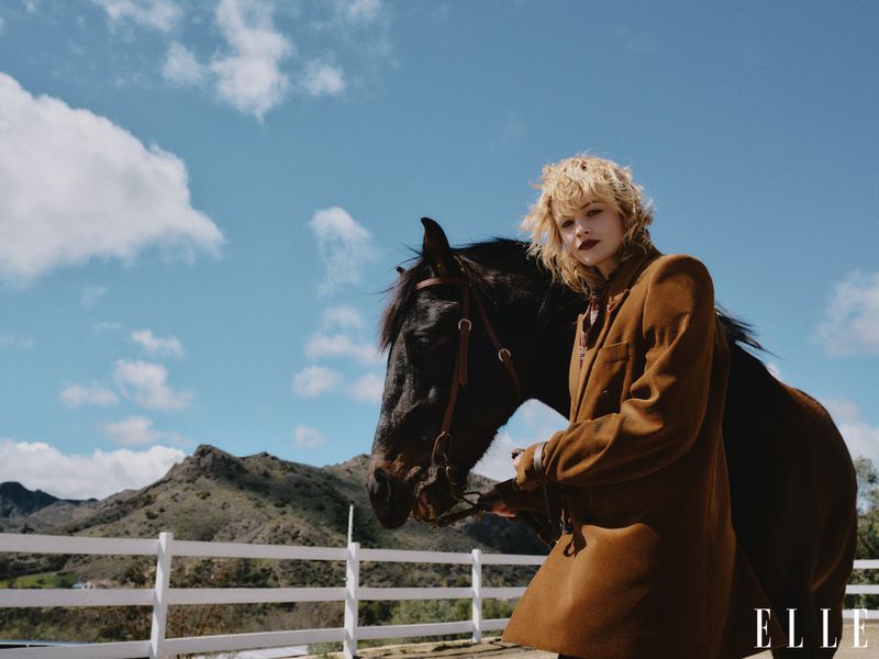 Model poses with a black horse outdoors in a brown coat, against a backdrop of rolling hills and a blue sky with scattered clouds.