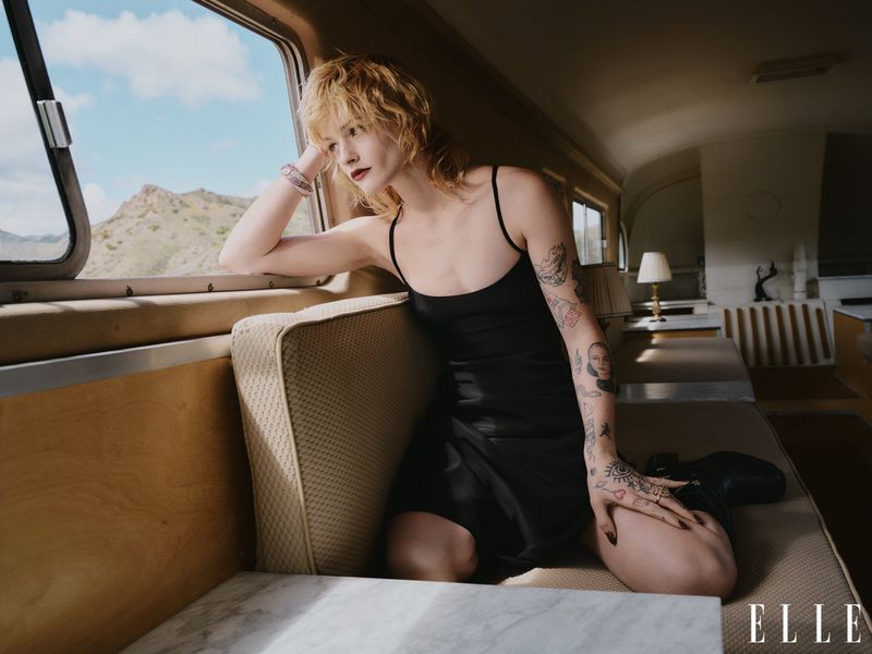 Woman in black dress looks out train window at mountains. Interior with benches, tables, lighting fixtures. Elle magazine logo visible.