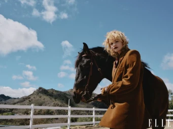 Blonde woman wearing a brown coat standing beside a black horse with a mountain range and white fence in the background.