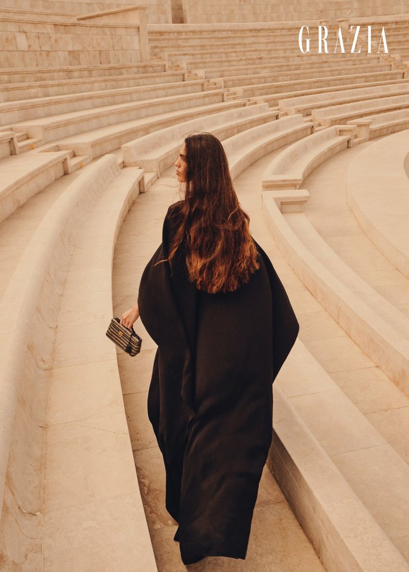 A woman in a black dress walks on the stone steps of an outdoor amphitheater, carrying a purse. Editorial fashion photograph with magazine logo.