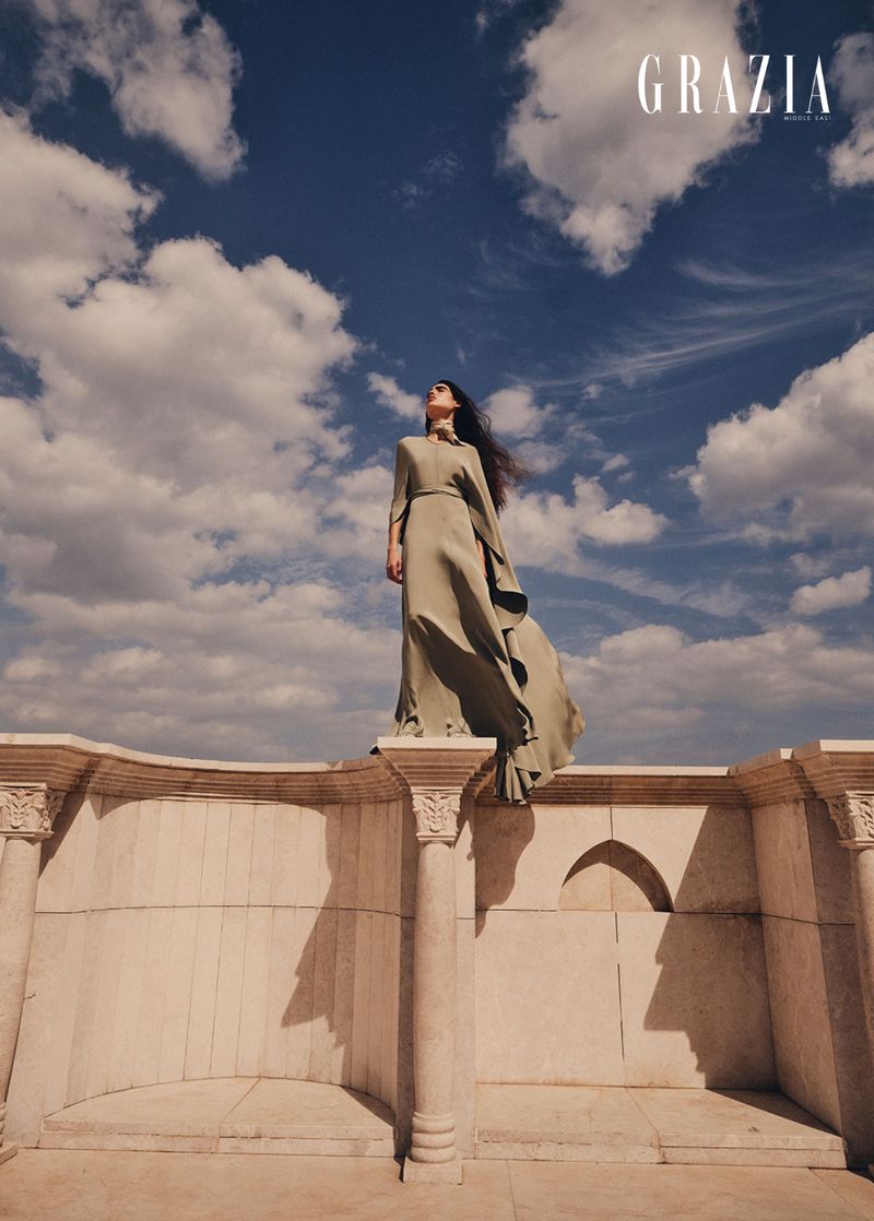 Fashion editorial shot of a woman in a long olive dress on a stone terrace against a bright blue sky with scattered clouds.