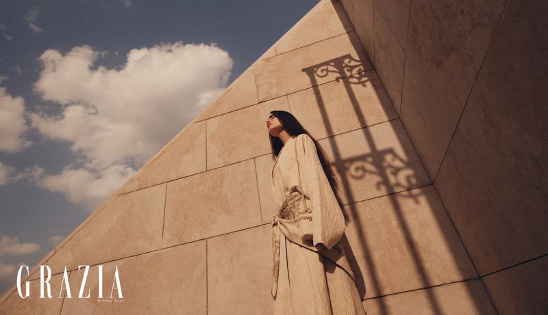Adult woman poses against an angular beige building under a blue sky with clouds in a fashion editorial shot for Grazia Middle East.