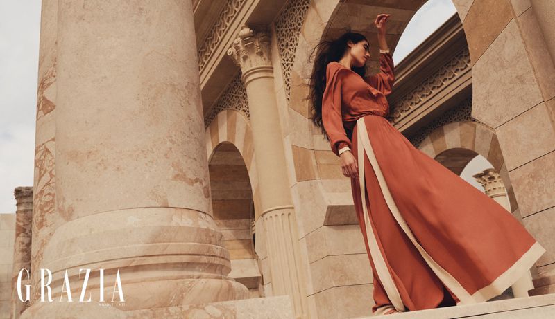 A woman models an earth-toned dress in front of sandstone arches and columns. The image features earth tones and architectural elements.