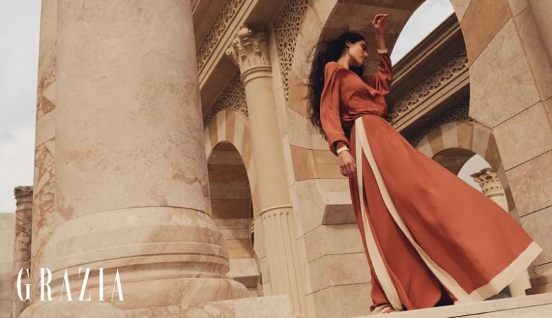 A woman models an earth-toned dress in front of sandstone arches and columns. The image features earth tones and architectural elements.