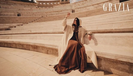 Model wearing a hooded coat and brown skirt seated in a grand stone amphitheater under natural light.