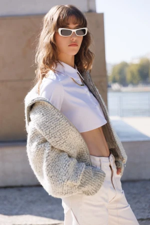 A young woman with curly brown hair poses outdoors wearing a white crop top, off-white pants, sunglasses, and a fuzzy gray cardigan.