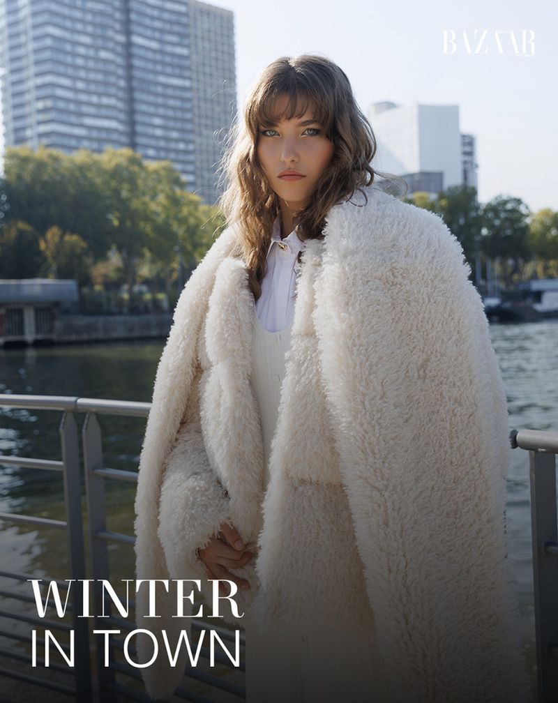 Fashion editorial shot of a woman with bangs in a cream, textured coat standing in front of an urban river scene on a sunny day.