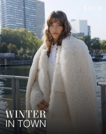 Fashion editorial shot of a woman with bangs in a cream, textured coat standing in front of an urban river scene on a sunny day.