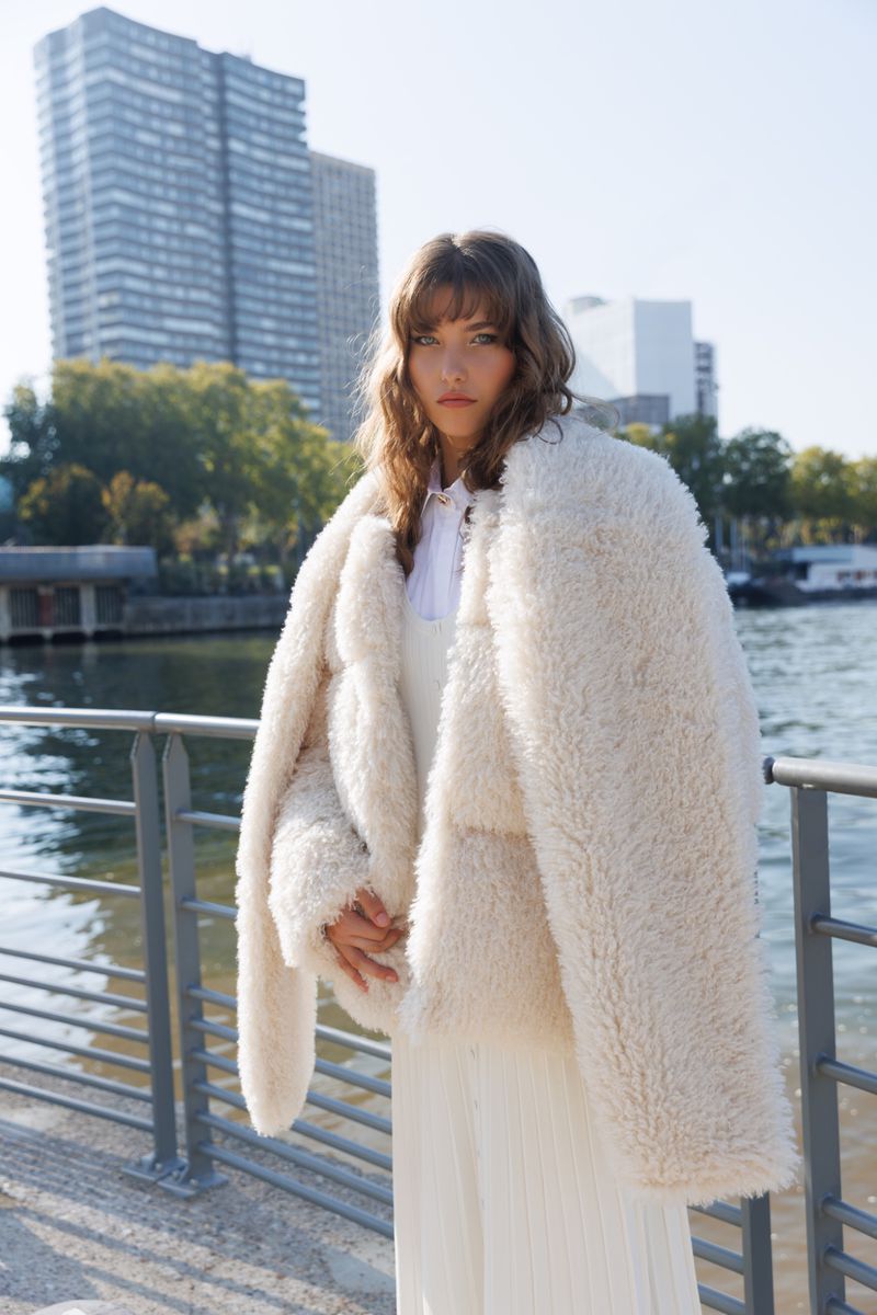 A young woman poses outdoors wearing an oversized, textured faux fur jacket with skyscrapers and a body of water in the background.